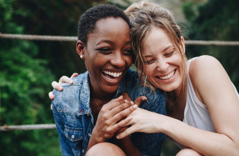 Two women smiling and laughing together outdoors, sitting closely with arms around each other. Green foliage is visible in the background.