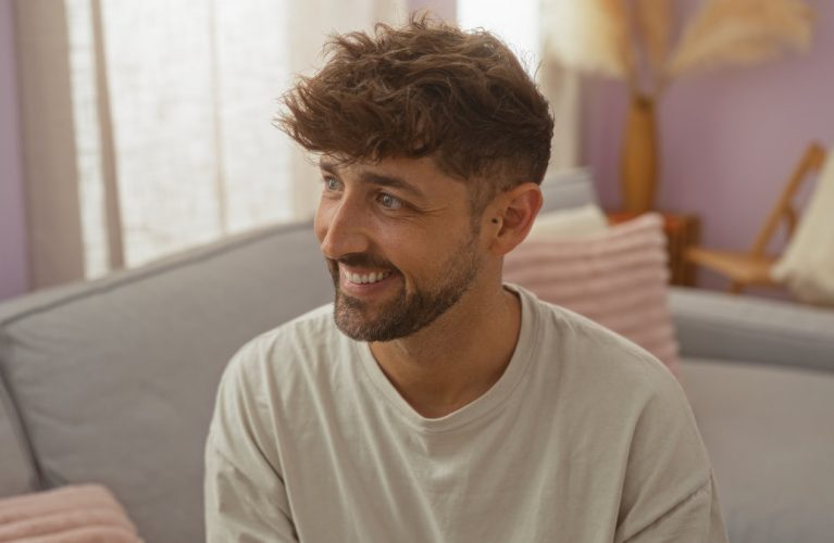 A man with short, wavy brown hair and a beard smiles while sitting on a light-colored couch in a softly lit living room.