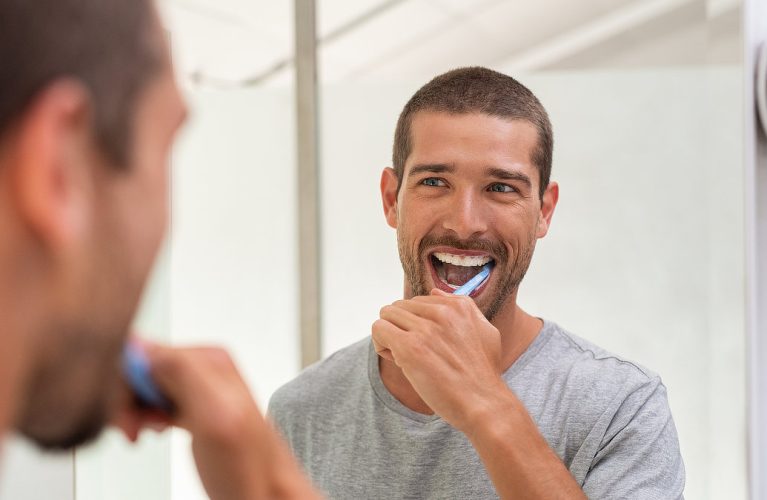 A man brushing his teeth in front of a bathroom mirror, wearing a gray t-shirt.