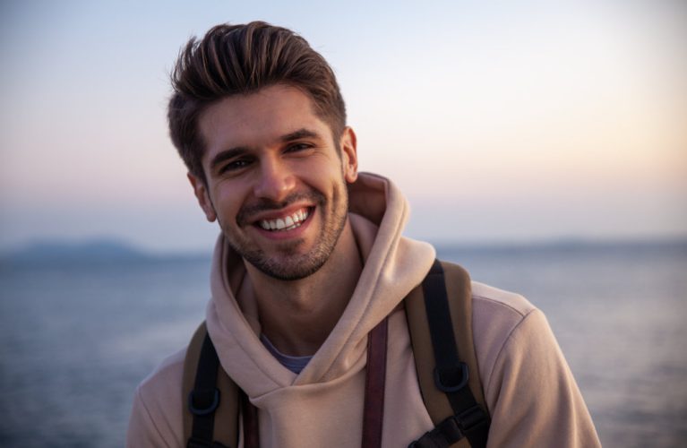 A young man wearing a beige hoodie and backpack smiles outdoors near a body of water at sunset.