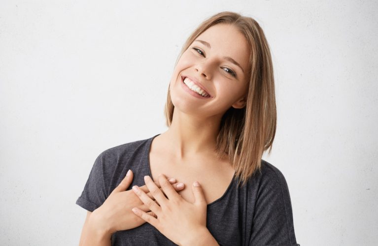 Beautiful positive friendly-looking young mixed race woman with lovely sincere smile feeling thankful and grateful, showing her heart filled with love and gratitude holding hands on her breast